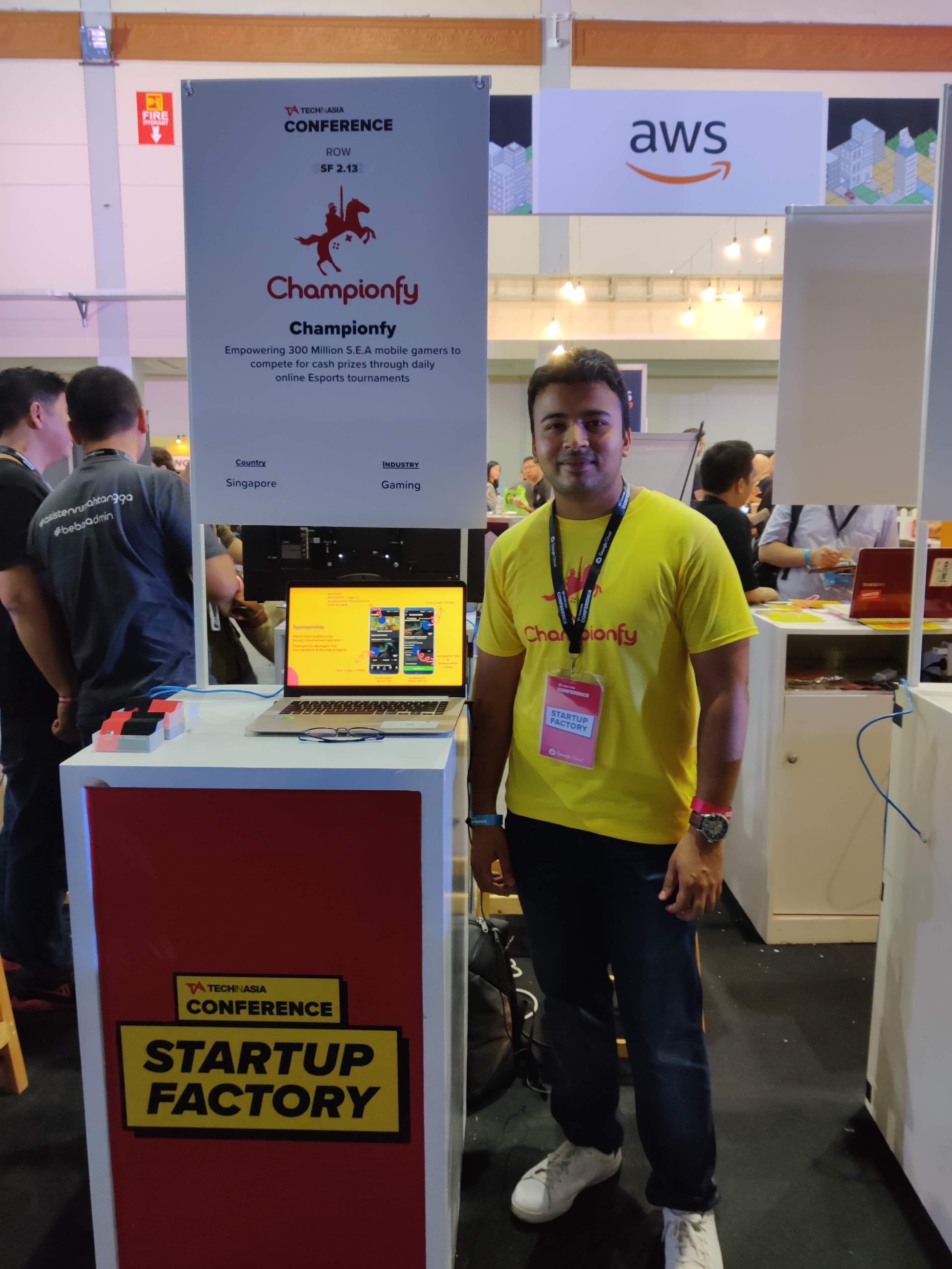 Standing at the Championfy booth in a yellow Championfy t-shirt, a laptop on the pod showing the pitch deck